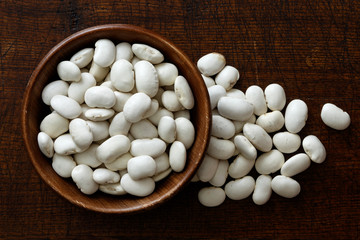 Fototapeta na wymiar Dry butter beans in dark wooden bowl isolated on dark brown wood from above. Spilled beans.