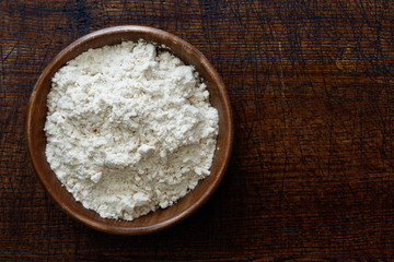 Fototapeta na wymiar Fine white flour in dark wooden bowl isolated on dark brown wood from above.