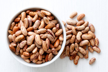 Dry pinto beans in white ceramic bowl isolated on painted white wood from above. Spilled beans.