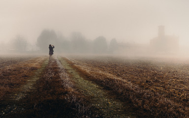 Unidentifable woman in the distance looking at a church in the dense fog of the Lombardy region of Italy during the winter, frozen corn field - obrazy, fototapety, plakaty