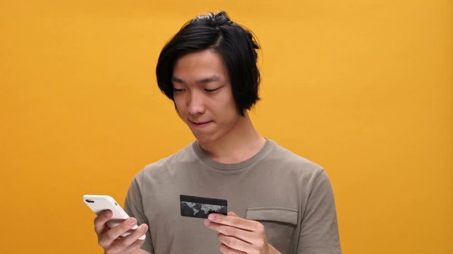 Young asian man smiling holding credit card using phone isolated over yellow background