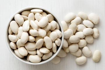 Dry butter beans in white ceranic bowl isolated on painted white wood from above. Spilled beans.