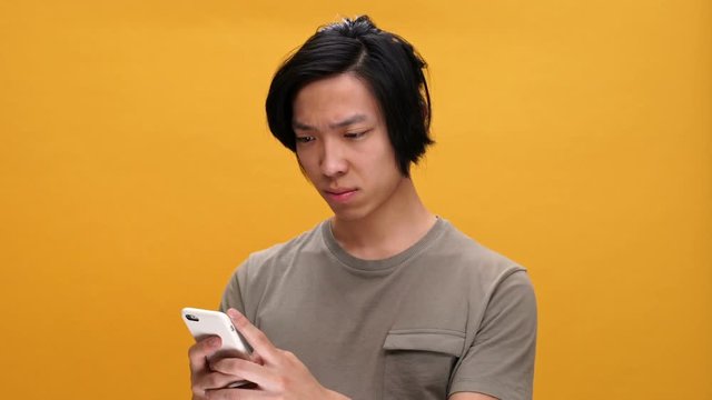 Young sad asian man chatting by phone isolated over yellow background