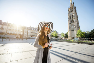 Young woman tourist walking with photocamera on the Pey-Berland square with bell tower on the background during the morning in Bordeaux city - obrazy, fototapety, plakaty