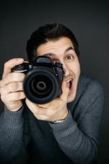 Fototapeta premium Young male photographer looking at the camera lens and smile. Funny expression. Studio portrait isolated on gray