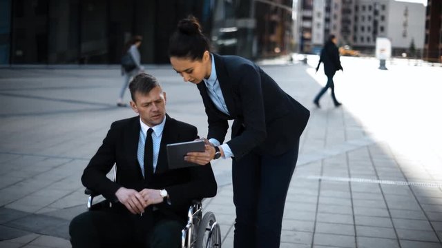 Invalid cripple boss businessman successful and powerful in wheelchair in suit and his lady secretary businesswaman working with digital pad, or tablet PC outdoor.