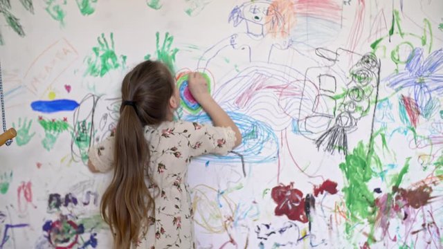 Medium shot with rear view of little girl in linen dress drawing colorful heart on wall of her room covered in doodles