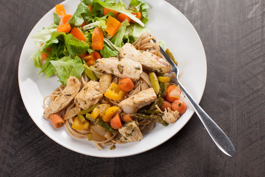  
Whole wheat pasta with cooked chicken and vegetables including carrots, bell peppers, onion, and asparagus with a small salad as a side slightly above shot
