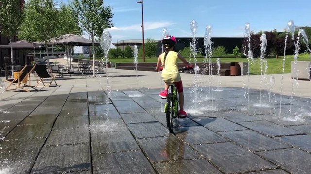 Little girls ride their bikes among water fountains in  hot summer day