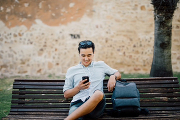 Happy asian guy using a smart phone sitting on a bench in a park
