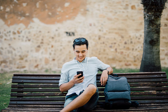 Happy Asian Guy Using A Smart Phone Sitting On A Bench In A Park