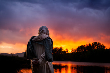 Jesus Christ stands beside a lake and watches the sun sets over the water.