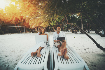 Two beautiful adult people on resort beach chairs: smiling handsome man watching movie via digital...