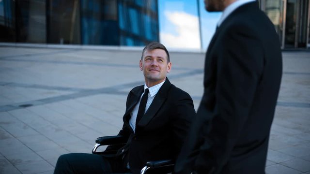 Business people in formal suits have conversation with the invalid man in wheelchair outdoor near buisness centre modern building, Handed shot. Tolerance concept. Teal and orange Sunrise time.