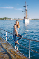 Portrait of white Caucasian blonde woman with tanned skin striped t-shirt and blue jeans sitting on pier by seashore lakeshore, with yacht boat ship on background on water, lifestyle summer