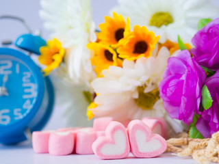 Fototapeta na wymiar Closeup of sweet marshmallow in the shape of heart and Alarm clock on wooden plate and flower at background. Concept about love and relationship. (Soft Style for Background)