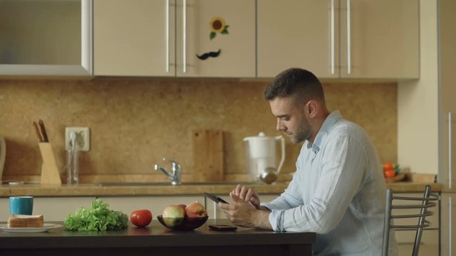 Serious young man using digital tablet computer sitting in the kitchen while have breakfast in the morning