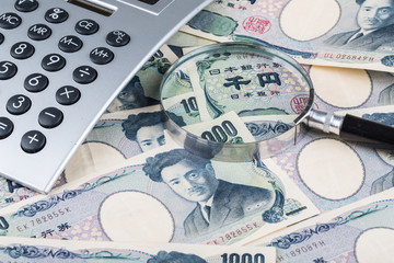 Fototapeta premium Stack of Japanese currency yen or Japanese banknotes and Japanese yen coins with magnifying glass and calculator