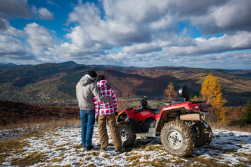 Rear view of a guy hugging a girl near the ATV on a mountain top under the blue sky with cumulus clouds at background of mighty mountains and village in the valley © anatoliy_gleb