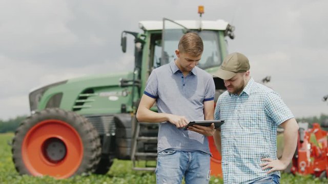 Two young men of the farmer communicate, use the tablet. Stand in the field against a tractor background