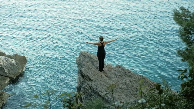 Woman on a rock by the beautiful pond raises his hands. She stands on a pendant stone and does stretching gymnastics.