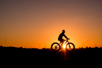 Naklejka na ściany i meble Silhouette of cyclist riding on a bike on road at sunset.