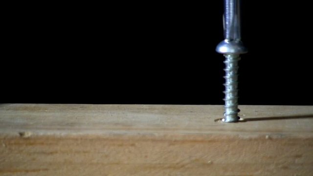 A pan head screw is tightened in pine board by hand with phillips head screwdriver on right side.  Macro shot straight on with black background.