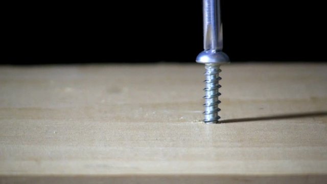 A pan head screw is tightened in pine board by hand with phillips head screwdriver on right side.  Macro shot from slight high angle looking down with black background.
