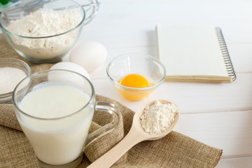 Baking in the kitchen - the ingredients of the test recipe (eggs, flour, milk, sugar) on a white wooden table with the inscription on top. Background layout with free text space.