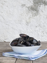 Fresh and raw sea mussels in white ceramic bowl resting on cotton blue and green napkin. Wooden table and white wall background. Side view.