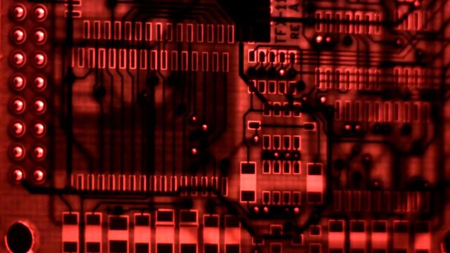 A single bare circuit board rotates on end from right to left with red back light revealing inner trace layers and component pads.  Light shines through holes and vias