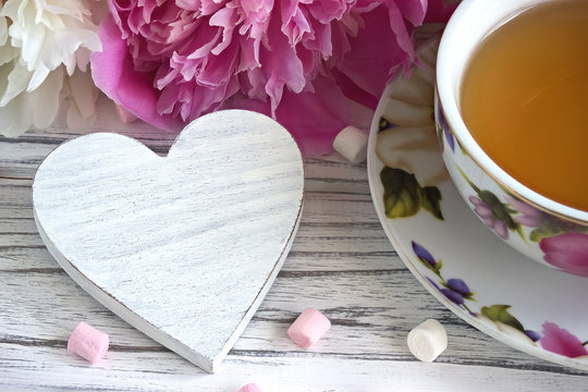 Peonies flowers pink cup of tea with white wooden heart marshmallow on a white wooden background - stock image.