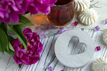 Peonies flowers pink glass of tea with white wooden heart marshmallow on a white wooden background - stock image.