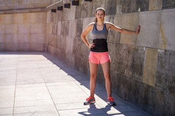 Fototapeta na wymiar Young smiling female resting after an active fitness training while standing against wall, standing in full sun