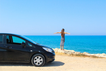 Fototapeta na wymiar A woman with her arms up next to a rented car with a view of the sea