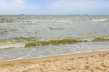 Seascape with ship in the distance and cloudy sky. Soft sea ocean waves wash over golden sand background. Beautiful seascape with water splashes. Selective focus on the splash
