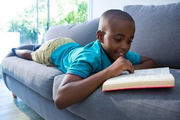 Smiling boy reading novel while lying on sofa at home