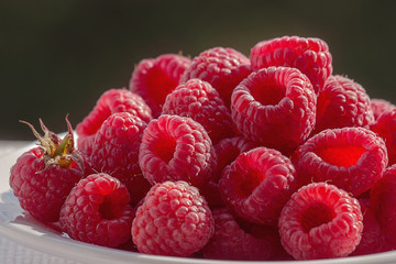 Raspberries on a plate closeup. Large and delicious raspberries presented in the backlight on dark blurred background.