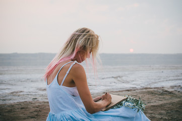 Fototapeta na wymiar Young hipster girl with pink hair sitting and drawing with pencil at the coast