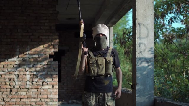 Male terrorist covering face standing with gun up and looking at camera in ruined building.