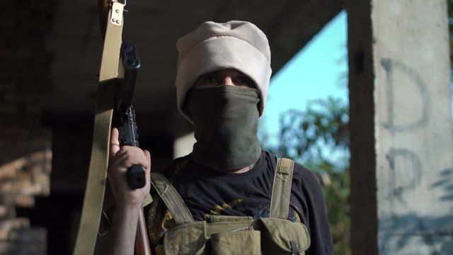 Terrorist in camouflage guarding a building with a bandanna around his face and cap pulled low over his eyes holding an automatic weapon looking at camera.