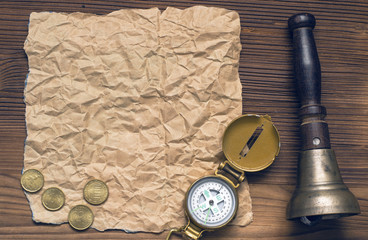 Compass, train bell, money and blank crumpled brown page paper on wooden table. Adventure, treasure hunt, journey or travel concept.