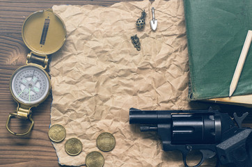 Adventurer, western, reward page, treasure hunt or travel concept. Compass, revolver gun, money, diary book, train conductor bell, pencil and blank crumpled brown page paper on wooden table.