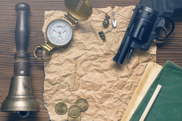 Adventurer, western, reward page, treasure hunt or travel concept. Compass, revolver gun, money, diary book, train conductor bell, pencil and blank crumpled brown page paper on wooden table.