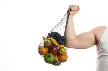 Athlete man flexing his bicep and holding a mesh shopping bag full of fruit isolated on white...