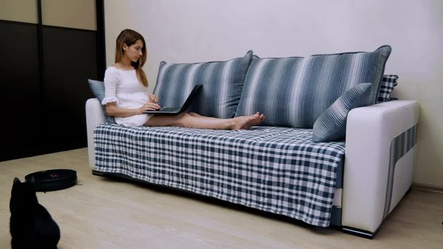 The young beautiful woman using a laptop on the sofa while the robotic vacuum cleaner is cleaning floor in the room. 4K