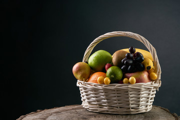 Assortment of fresh raw fruits in wicker basket on wooden stump isolated on black background. Healthy eating concept.