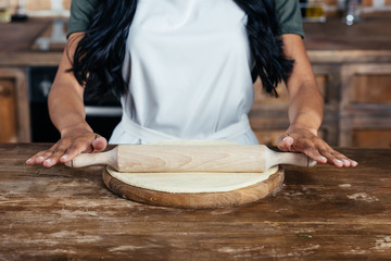 Cropped shot of woman in apron rolling pizza dough on wooden board