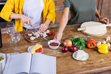 Cropped shot of young women cooking pizza while standing together at kitchen table with blank cookbook