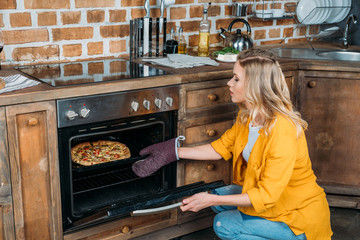 Beautiful young blonde woman baking pizza in oven in the kitchen